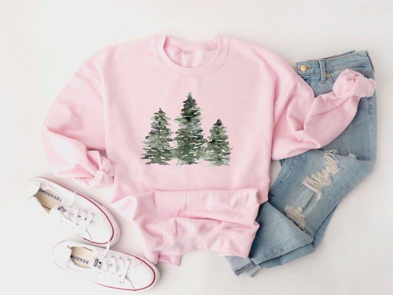 Puede incluir: Una sudadera rosa con un dise&ntilde;o de acuarela de tres &aacute;rboles verdes. La sudadera est&aacute; colocada sobre una superficie blanca con un par de zapatillas blancas y unos vaqueros azules.