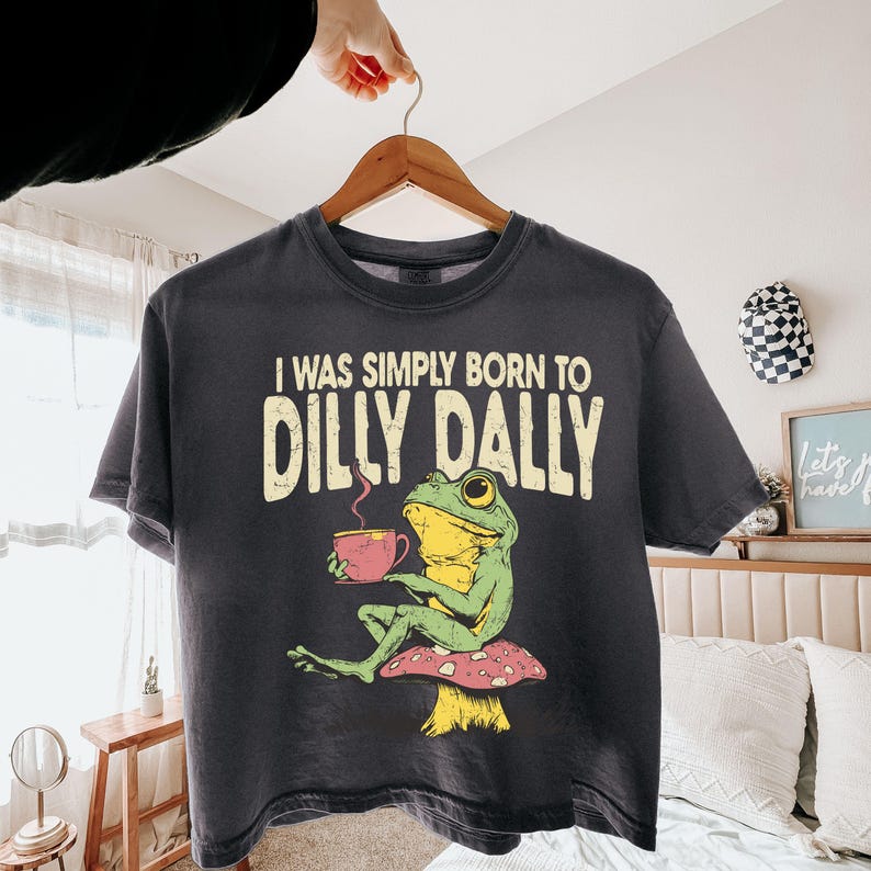 May include: Dark grey t-shirt with the text "I WAS SIMPLY BORN TO DILLY DALLY". The shirt features a cartoon frog sitting on a mushroom and holding a pink cup. The frog is green and yellow.