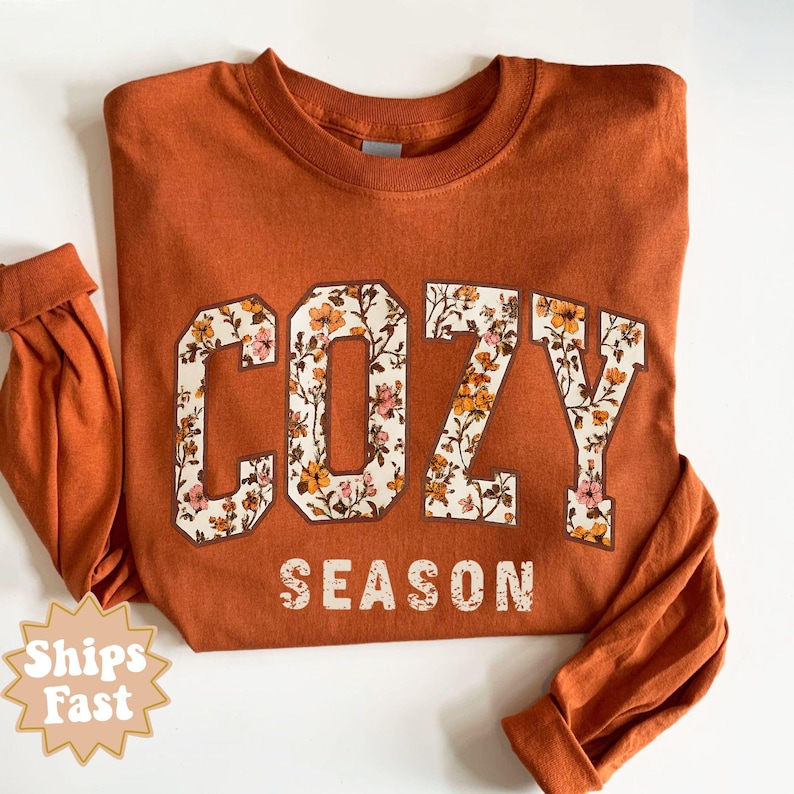 May include: An orange long-sleeve sweatshirt with the words "COZY SEASON" in a floral design. The letters are outlined in brown and filled with a pattern of flowers in shades of pink, orange, and white. The sleeves are cuffed.