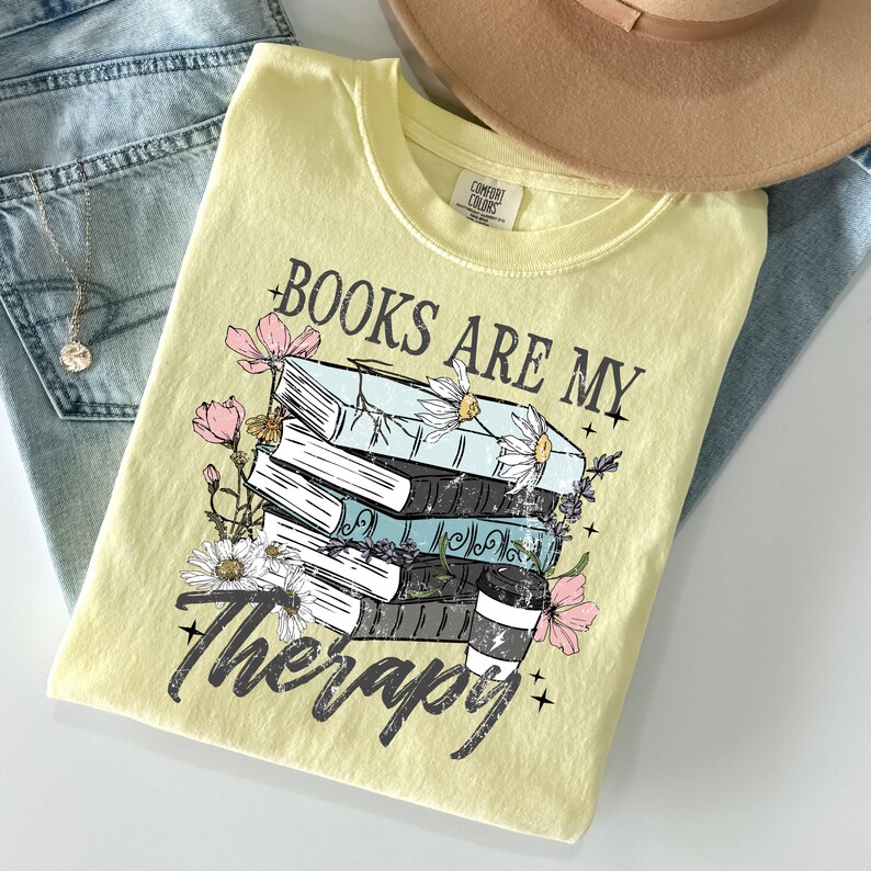 May include: A light yellow t-shirt with the text "BOOKS ARE MY Therapy" and a graphic of stacked books, flowers, and a coffee cup. The shirt is folded on top of blue jeans and a tan hat.