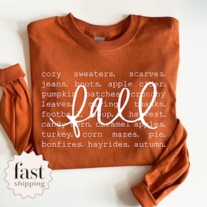 May include: A burnt orange long-sleeve crewneck shirt featuring white text listing fall-themed words including cozy sweaters, scarves, jeans, boots, apple cider, pumpkin patches, crunchy leaves, giving thanks, football, soup, harvest, candy corn, caramel apples, turkey, corn mazes, pie, bonfires, hayrides, and autumn.