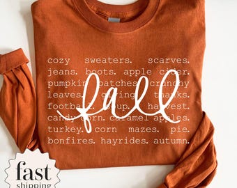 Puede incluir: Una camiseta de manga larga y cuello redondo en color naranja quemado con texto blanco que enumera palabras relacionadas con el otoño, incluyendo cozy sweaters, scarves, jeans, boots, apple cider, pumpkin patches, crunchy leaves, giving thanks, football, soup, harvest, candy corn, caramel apples, turkey, corn mazes, pie, bonfires, hayrides y autumn.
