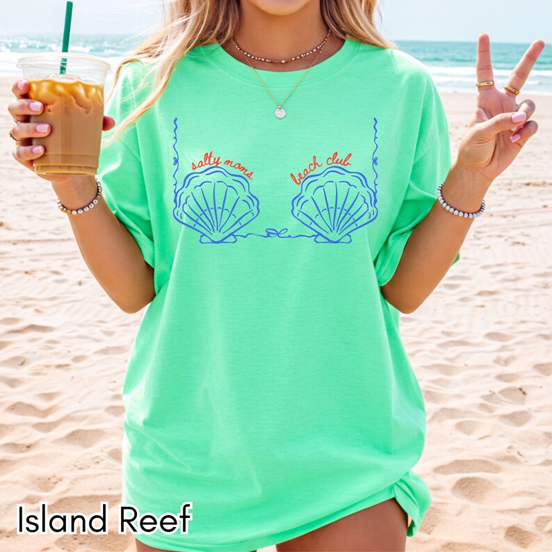 May include: A light green t-shirt with the text "salty moms beach club" above a blue seashell design. The words "Island Reef" are printed at the bottom of the shirt.