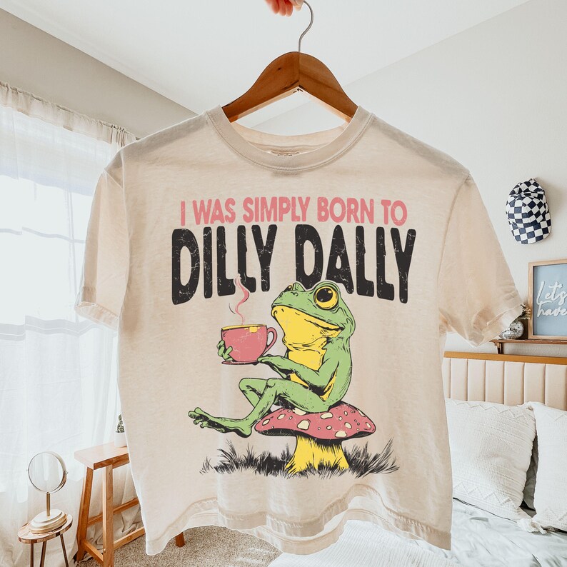 May include: A cream-coloured t-shirt with a frog illustration. The frog, in green and yellow, sits on a mushroom and holds a pink cup. The shirt has the text "I WAS SIMPLY BORN TO DILLY DALLY" in black and pink.