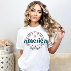 May include: White t-shirt with a patriotic design. The design features the word "America" in blue, encircled by a red ring with the text "Land of the Free" and "Because of the Brave." A peace sign is included.