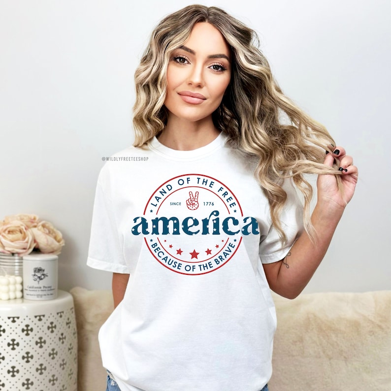 May include: White t-shirt with a patriotic design. The design features the word "America" in blue, encircled by a red ring with the text "Land of the Free" and "Because of the Brave." A peace sign is included.