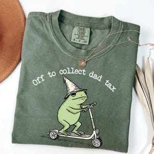 May include: Olive green t-shirt with the phrase "Off to collect dad tax" and a cartoon frog riding a scooter. The frog wears a wizard hat. A gold necklace is draped across the shirt.