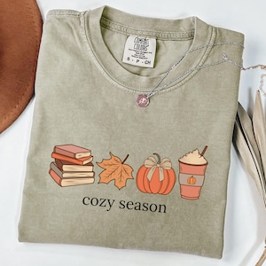 May include: Sage green t-shirt featuring a fall graphic with a stack of books, a maple leaf, a pumpkin, and a coffee cup, with the words "cozy season". A silver necklace is draped across the shirt.