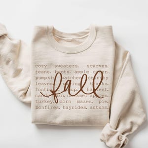 Pode incluir: Uma camisola de gola redonda cor de creme com texto castanho listando palavras com o tema de outono, incluindo cozy sweaters, scarves, jeans, boots, apple cider, pumpkin patches, crunchy leaves, giving thanks, football, soup, harvest, candy corn, caramel apples, turkey, corn mazes, pie, bonfires, hayrides, autumn, com a palavra fall escrita em letra cursiva grande.