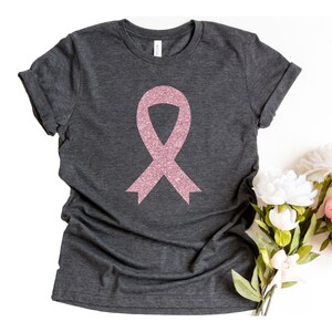 May include: A dark charcoal grey short-sleeve t-shirt featuring a centred pink glitter awareness ribbon graphic. The shirt has a crew neckline and rolled sleeves, showcasing a textured fabric finish with a visible brand label at the inner collar.