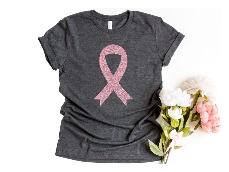 May include: A dark charcoal grey short-sleeve t-shirt featuring a centred pink glitter awareness ribbon graphic. The shirt has a crew neckline and rolled sleeves, showcasing a textured fabric finish with a visible brand label at the inner collar.