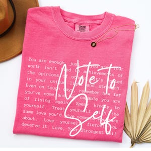 Może przedstawiać: Różowa koszulka Comfort Colors z napisem "Note to Self" w białym skrypcie. Koszulka ma okrągły dekolt i zawiera inspirujący tekst. Złoty naszyjnik z czarnym wisiorkiem jest zarzucony na koszulkę.