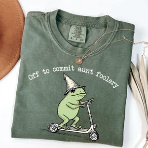 May include: Sage green t-shirt with the phrase "Off to commit aunt foolery" and a cartoon frog riding a scooter. The frog wears a wizard hat. A gold necklace and a brown hat are also visible.