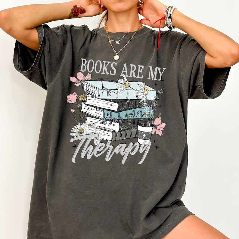 May include: Dark gray t-shirt with the text "BOOKS ARE MY Therapy" in white script. The graphic features a stack of books with floral accents in pink, white, and blue. A coffee cup is also included in the design.