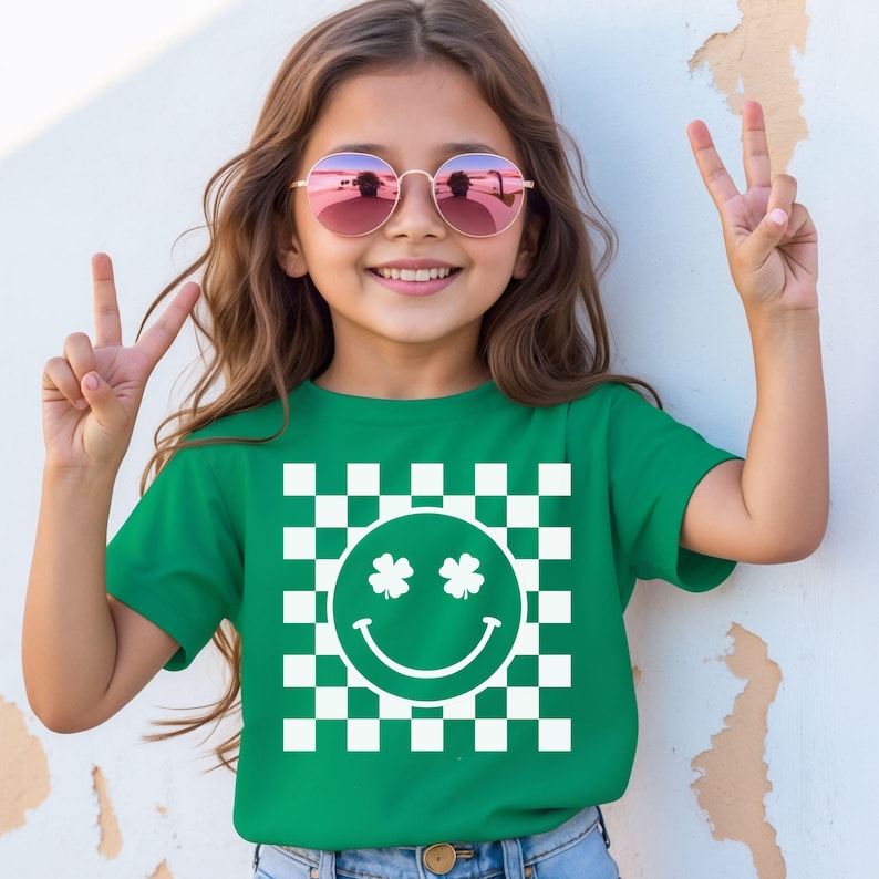 May include: A green t-shirt featuring a white checkered pattern with a smiling face and two shamrocks. The girl is wearing pink sunglasses and making a peace sign with her fingers.