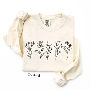 May include: Ivory-colored long-sleeve shirt with a crew neck. The shirt features a black line drawing of various flowers across the front. The word "Ivory" is printed in black at the bottom of the shirt.