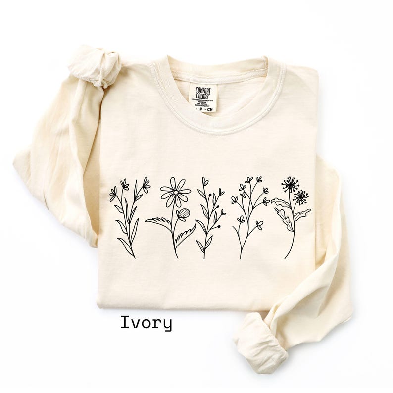 May include: Ivory-colored long-sleeve shirt with a crew neck. The shirt features a black line drawing of various flowers across the front. The word "Ivory" is printed in black at the bottom of the shirt.