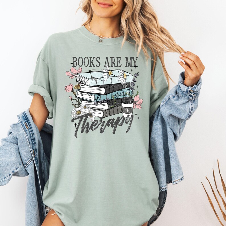 May include: A light green t-shirt with the text "BOOKS ARE MY Therapy" and a graphic of stacked books, flowers, and a coffee cup. The design is in black, white, pink, and blue. The shirt has short sleeves and a crew neck.