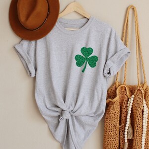 May include: A gray t-shirt with a green glitter shamrock design. The shirt is tied at the front and is hanging on a wooden hanger.