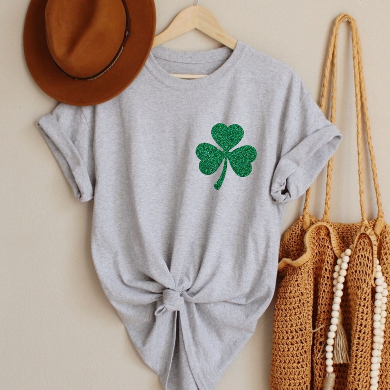 May include: A gray t-shirt with a green glitter shamrock design. The shirt is tied at the front and is hanging on a wooden hanger.