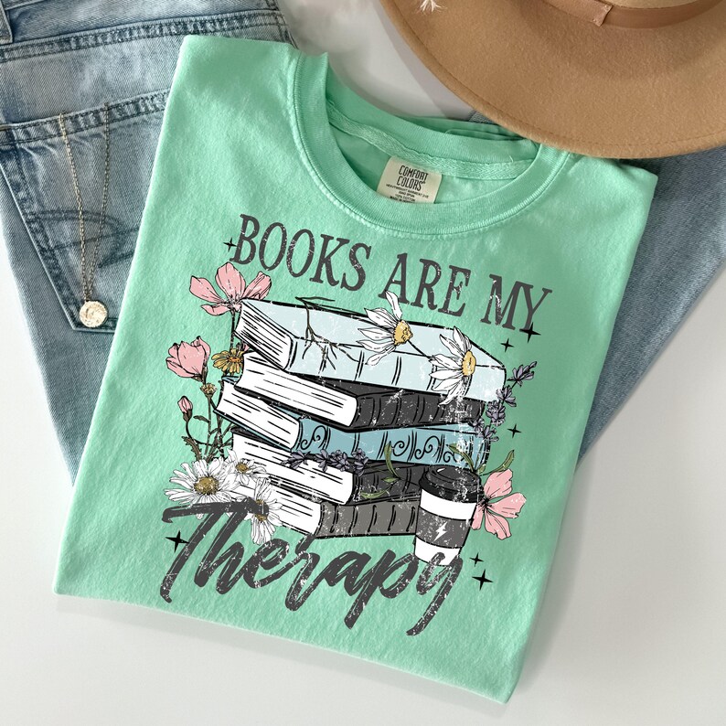 May include: A light green t-shirt with the text "BOOKS ARE MY Therapy" and a graphic of stacked books, flowers, and a coffee cup. The shirt is laid on top of blue jeans and a tan hat.