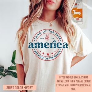 May include: Ivory-colored t-shirt with a patriotic design. The design includes the word "America" in blue, surrounded by a red circle with the text "Land of the Free Since 1776" and "Because of the Brave".