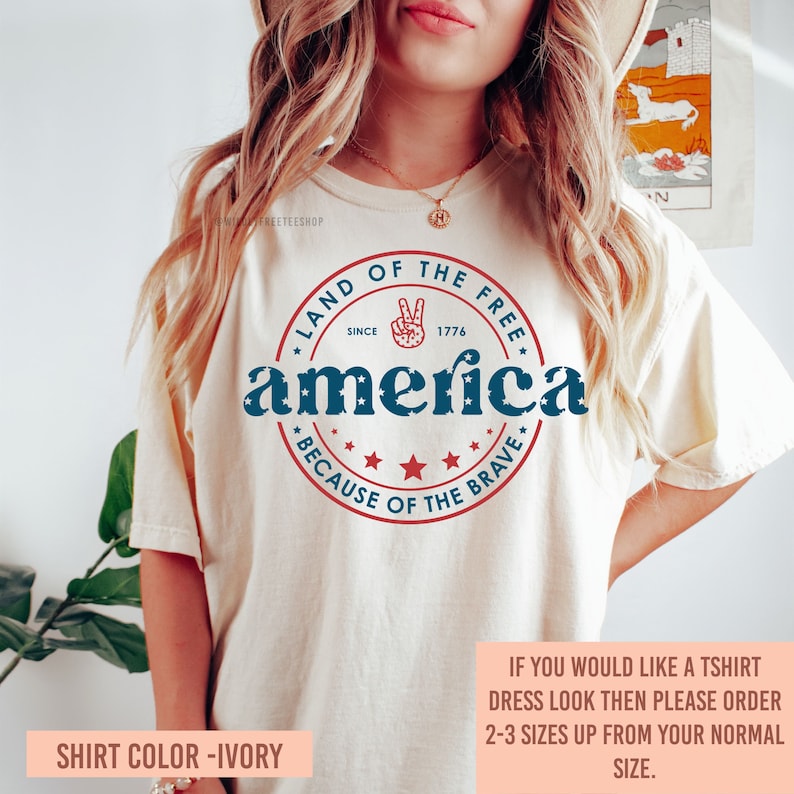 May include: Ivory-colored t-shirt with a patriotic design. The design includes the word "America" in blue, surrounded by a red circle with the text "Land of the Free Since 1776" and "Because of the Brave".