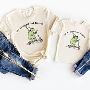May include: Two cream-colored t-shirts with the text "Off to commit dad foolery" and "Off to commit son foolery". Each shirt features a green frog riding a scooter. Blue jeans and dried flowers are also in the image.