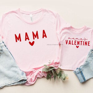 May include: Two pink t-shirts with red text. The larger shirt says "MAMA" with a red heart. The smaller shirt says "mama's VALENTINE" with a red heart.