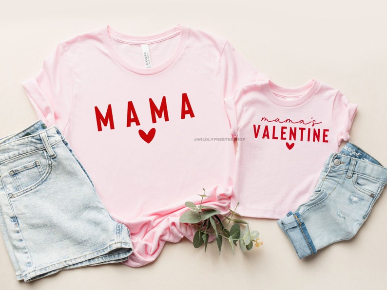 May include: Two pink t-shirts with red text. The larger shirt says "MAMA" with a red heart. The smaller shirt says "mama's VALENTINE" with a red heart.
