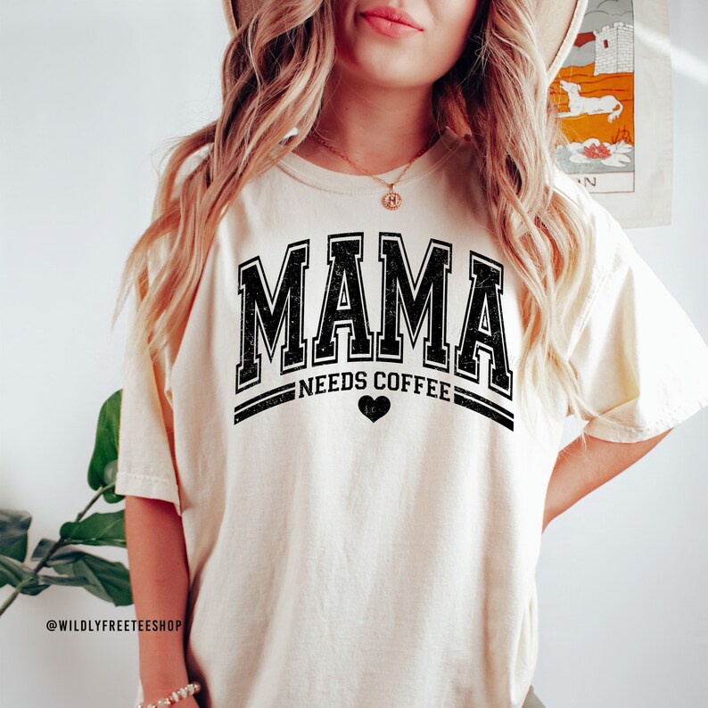 May include: A cream-coloured short-sleeve t-shirt featuring the black text MAMA in a distressed varsity font, with the words NEEDS COFFEE and a small heart icon centred below. The design is printed on the front of the garment.