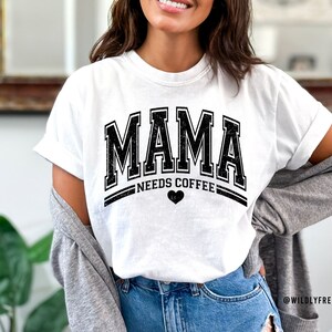 May include: A white short-sleeved t-shirt featuring the black distressed text MAMA in a collegiate font, with the words NEEDS COFFEE underneath and a small black heart icon. The shirt is worn with a grey cardigan and blue denim jeans.