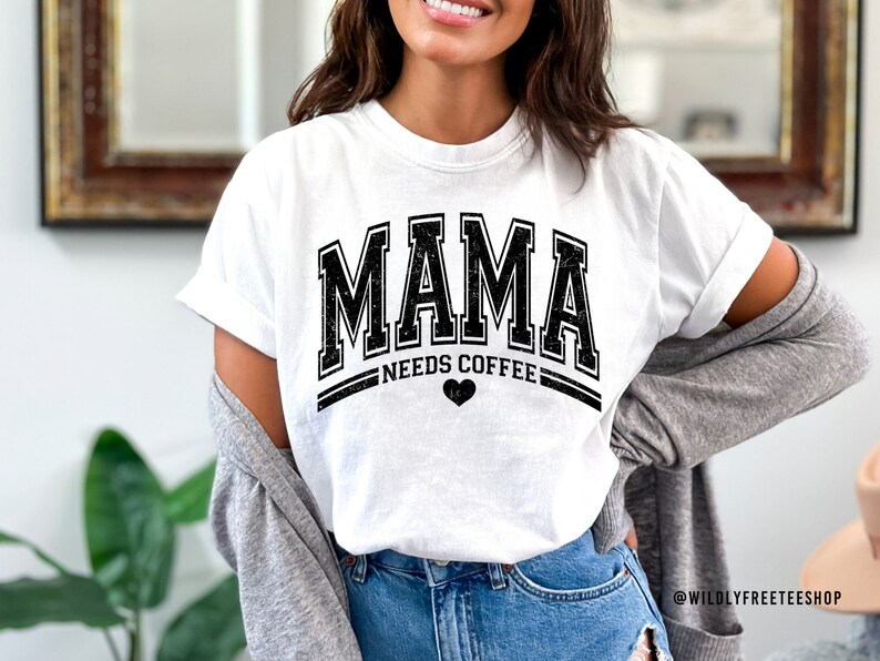 May include: A white short-sleeved t-shirt featuring the black distressed text MAMA in a collegiate font, with the words NEEDS COFFEE underneath and a small black heart icon. The shirt is worn with a grey cardigan and blue denim jeans.