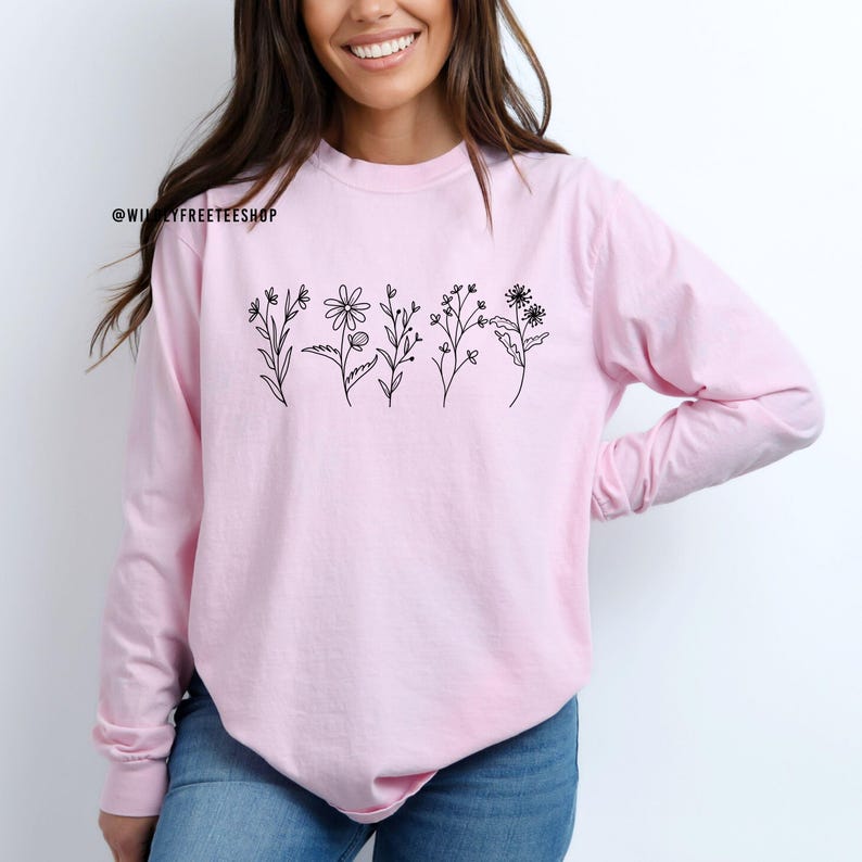 May include: A light pink long-sleeve shirt with a black floral design across the chest. The design features five different flower illustrations. The text "@WILYFREETEESHOP" is printed in black on the upper left side of the shirt.