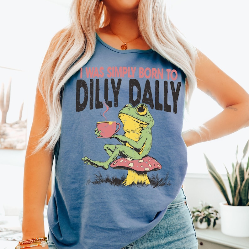 Op de afbeelding: Blauw tanktop met de tekst "I WAS SIMPLY BORN TO DILLY DALLY" en een afbeelding van een kikker op een paddenstoel, met een roze kopje. De kikker is groen en geel.