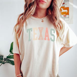 May include: A cream colored t-shirt with the word "TEXAS" printed in pastel green, yellow, pink, and blue.