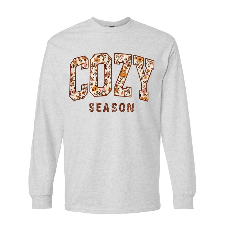 May include: A light gray long-sleeve shirt with the words "COZY SEASON" printed on the front. The word "COZY" is in a block letter style with a floral pattern. The word "SEASON" is in a brown, distressed font.