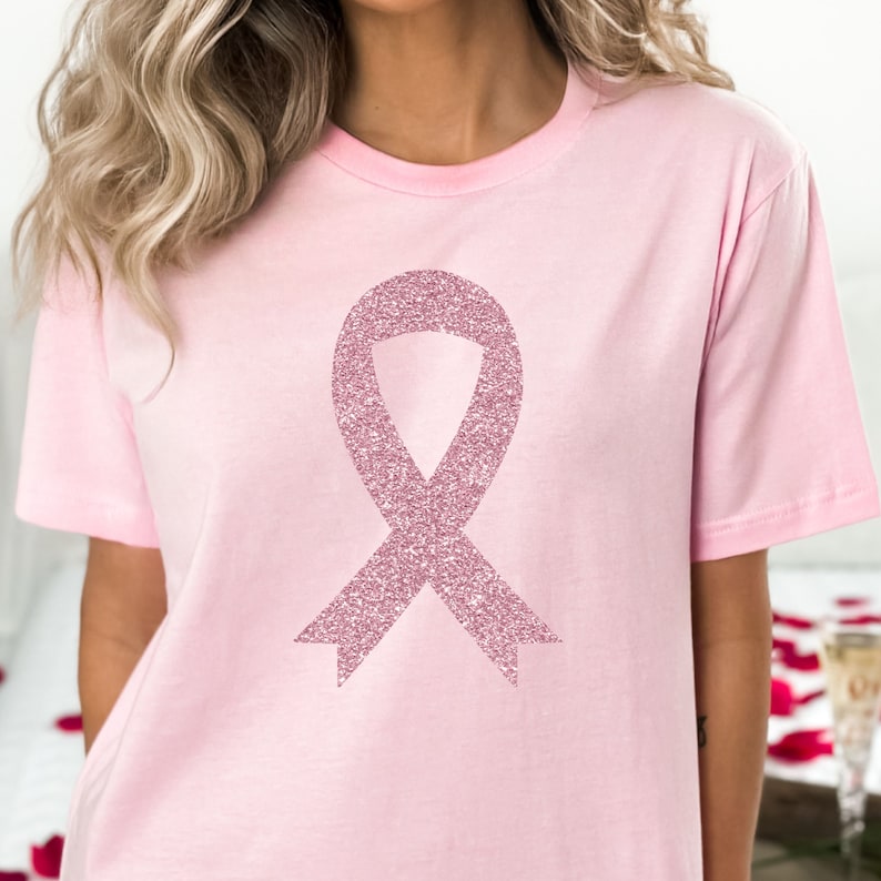 May include: A light pink short-sleeved t-shirt featuring a large, centred awareness ribbon graphic. The ribbon is rendered in a textured, sparkling rose-gold glitter finish that contrasts with the smooth, solid light pink fabric of the shirt.