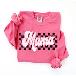 Puede incluir: Camiseta rosa de manga larga con un gr&aacute;fico blanco de "Mama" sobre un fondo a cuadros blanco y negro.