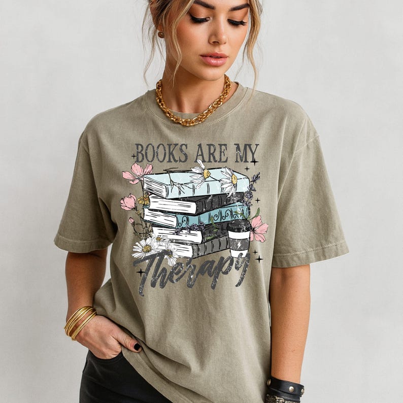 May include: Olive green t-shirt with the text "BOOKS ARE MY Therapy" and a graphic of stacked books, flowers, and a coffee cup. The design features light blue, white, and pink elements.