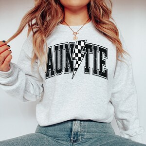 K&ouml;nnte beinhalten: Ein hellgraues Sweatshirt mit Rundhalsausschnitt und dem Wort AUNTIE in einer schwarzen, abgenutzten College-Schriftart. Der Buchstabe I ist durch einen schwarz-wei&szlig; karierten Blitz ersetzt. Das Design ist mittig auf der Brust platziert.
