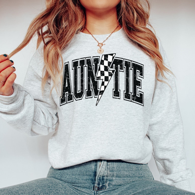K&ouml;nnte beinhalten: Ein hellgraues Sweatshirt mit Rundhalsausschnitt und dem Wort AUNTIE in einer schwarzen, abgenutzten College-Schriftart. Der Buchstabe I ist durch einen schwarz-wei&szlig; karierten Blitz ersetzt. Das Design ist mittig auf der Brust platziert.
