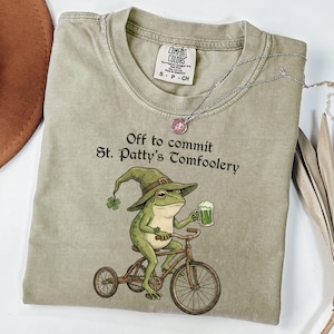 May include: A sage green t-shirt with the text "Off to commit St. Patty's Tomfoolery." A cartoon frog wearing a green hat and riding a bicycle is featured on the front. The frog is holding a mug of green beer.