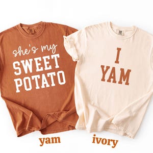 以下が含まれることがあります： 「she's my SWEET POTATO」と「I YAM」の文字がそれぞれ白と茶色でプリントされた、2枚のお揃いのTシャツ。色は、ヤムイモ色とアイボリー。白い背景に配置されています。
