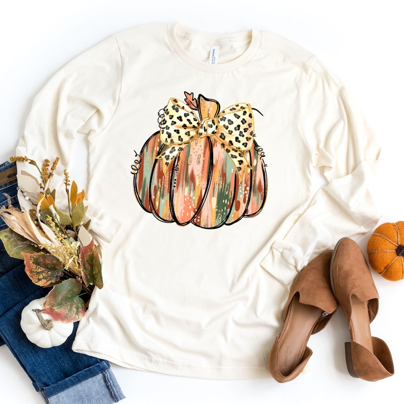 May include: Cream-colored long-sleeve shirt featuring a colorful pumpkin graphic with a leopard print bow. The pumpkin design includes shades of orange, green, and brown. The shirt is paired with jeans, fall-themed decor, and brown shoes.