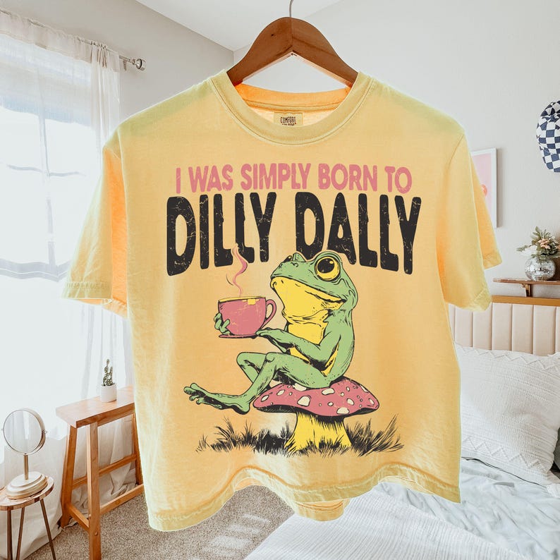 May include: Yellow t-shirt with the text "I WAS SIMPLY BORN TO DILLY DALLY" in black and pink. The shirt features a cartoon frog holding a pink teacup while sitting on a mushroom. The shirt is hanging on a wooden hanger.