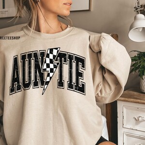 K&ouml;nnte beinhalten: Ein beigefarbenes Sweatshirt mit Rundhalsausschnitt und dem Wort AUNTIE in gro&szlig;en, schwarzen Blockbuchstaben mit Used-Optik. Der Buchstabe I ist durch einen schwarz-wei&szlig; karierten Blitz ersetzt. Der Text WILDLYFREETEESHOP ist in der Ecke zu sehen.