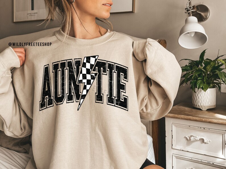 K&ouml;nnte beinhalten: Ein beigefarbenes Sweatshirt mit Rundhalsausschnitt und dem Wort AUNTIE in gro&szlig;en, schwarzen Blockbuchstaben mit Used-Optik. Der Buchstabe I ist durch einen schwarz-wei&szlig; karierten Blitz ersetzt. Der Text WILDLYFREETEESHOP ist in der Ecke zu sehen.