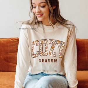 May include: Cream-colored long-sleeve shirt with the words "COZY SEASON" in a floral design. The shirt is made of a soft material and is perfect for fall. The shop's name, @WILDLYFREETEESHOP, is printed at the top.