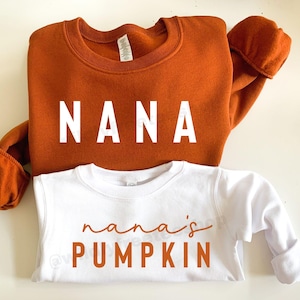 May include: An orange sweatshirt with the word "NANA" in white block letters is paired with a white long-sleeved shirt that says "nana's PUMPKIN" in orange script. The shirts are likely for a grandmother and grandchild.